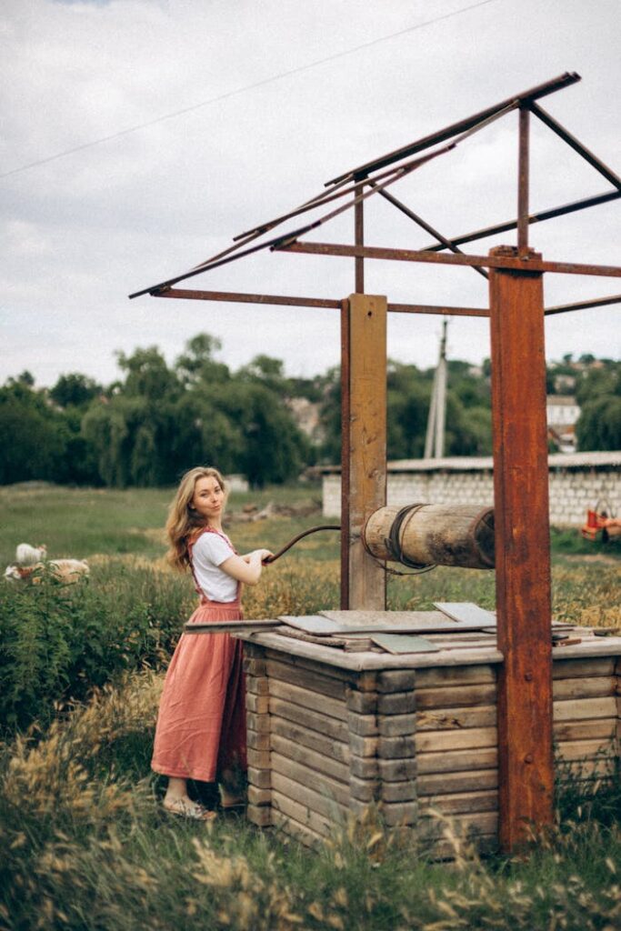 A young woman using a wooden well in a rural countryside setting with fields and trees.